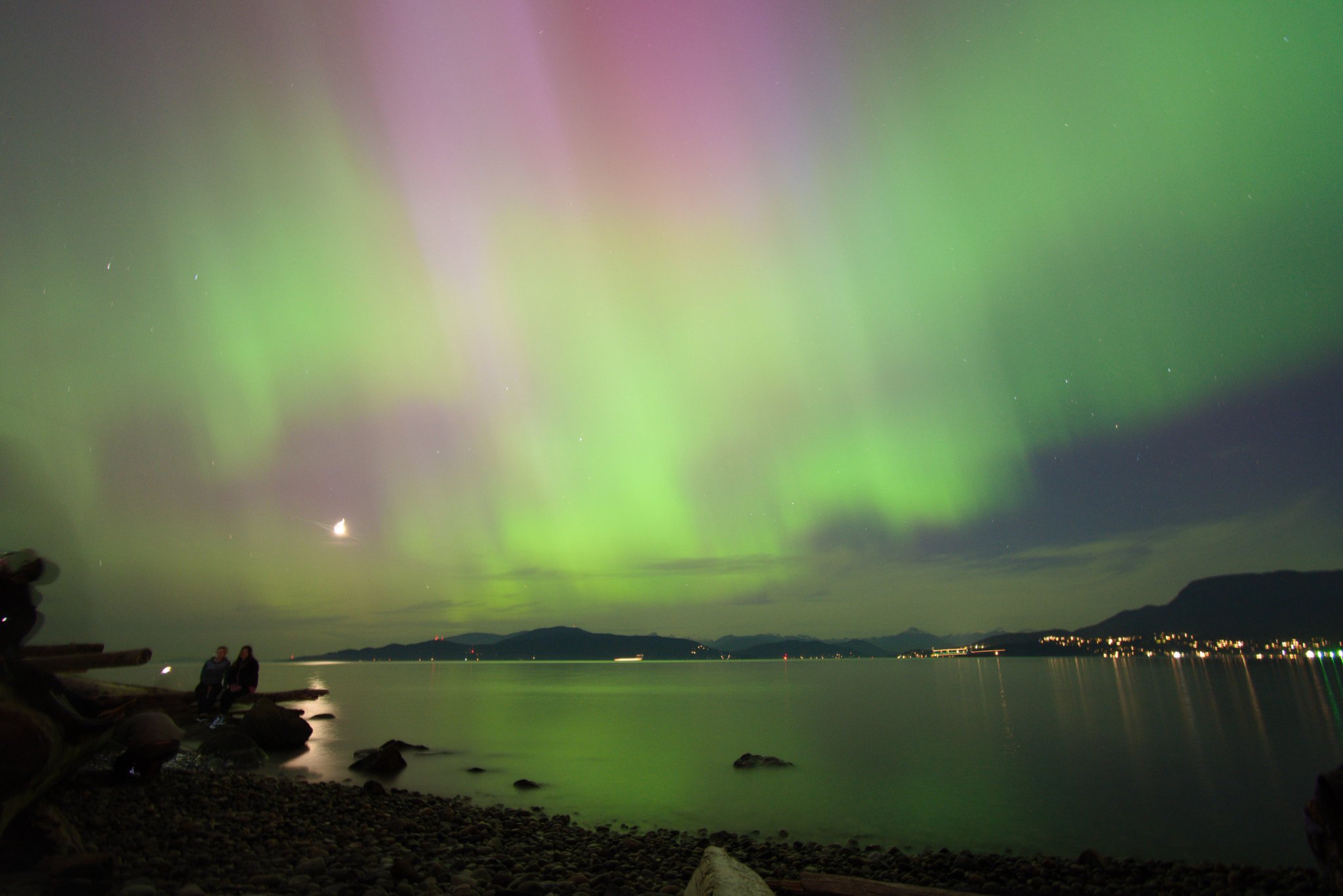 A picture of the Aurora Borealis, taken at a beach with rocky pebbles. To the right is some city lights, and to the left is something that vaguely resembles the moon. All lights can be seen reflecting onto the water.

Also there are people there.