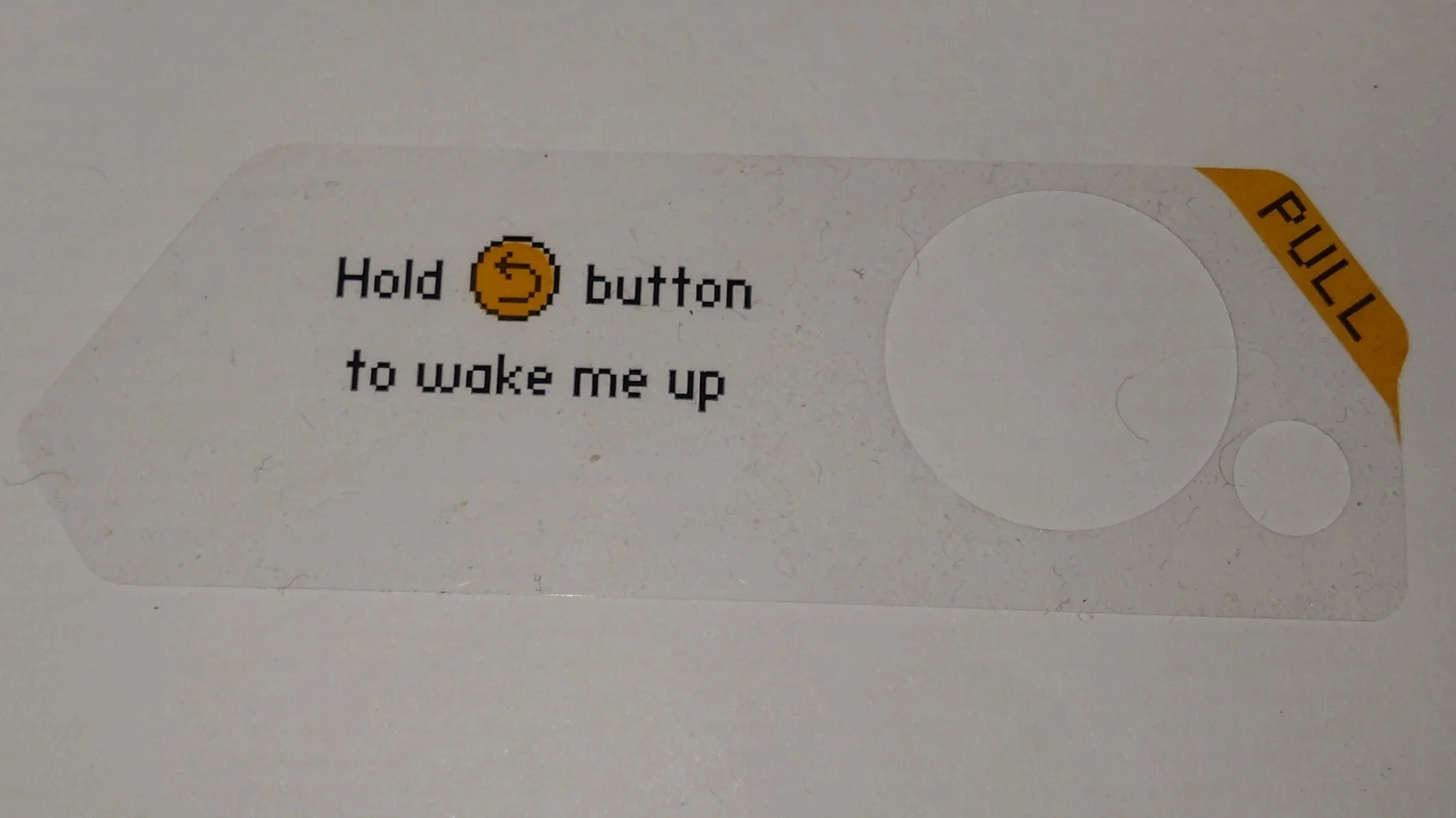 a transparent sticker vaguely in the shape of a flipper zero saying "Hold [icon of a back button] button to wake me up". The upper right corner says "pull".