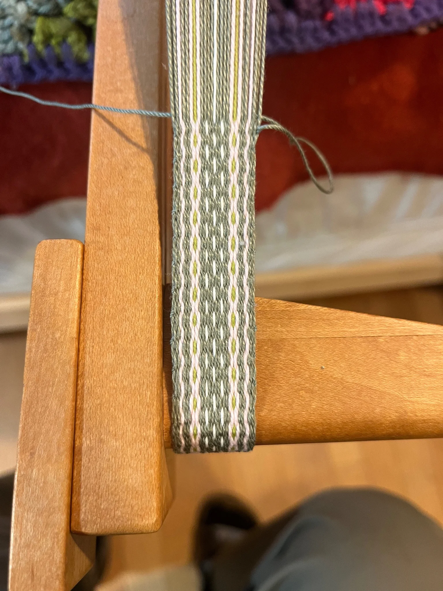 An olive green band with pale pink, lime green, and white details in the process of being woven