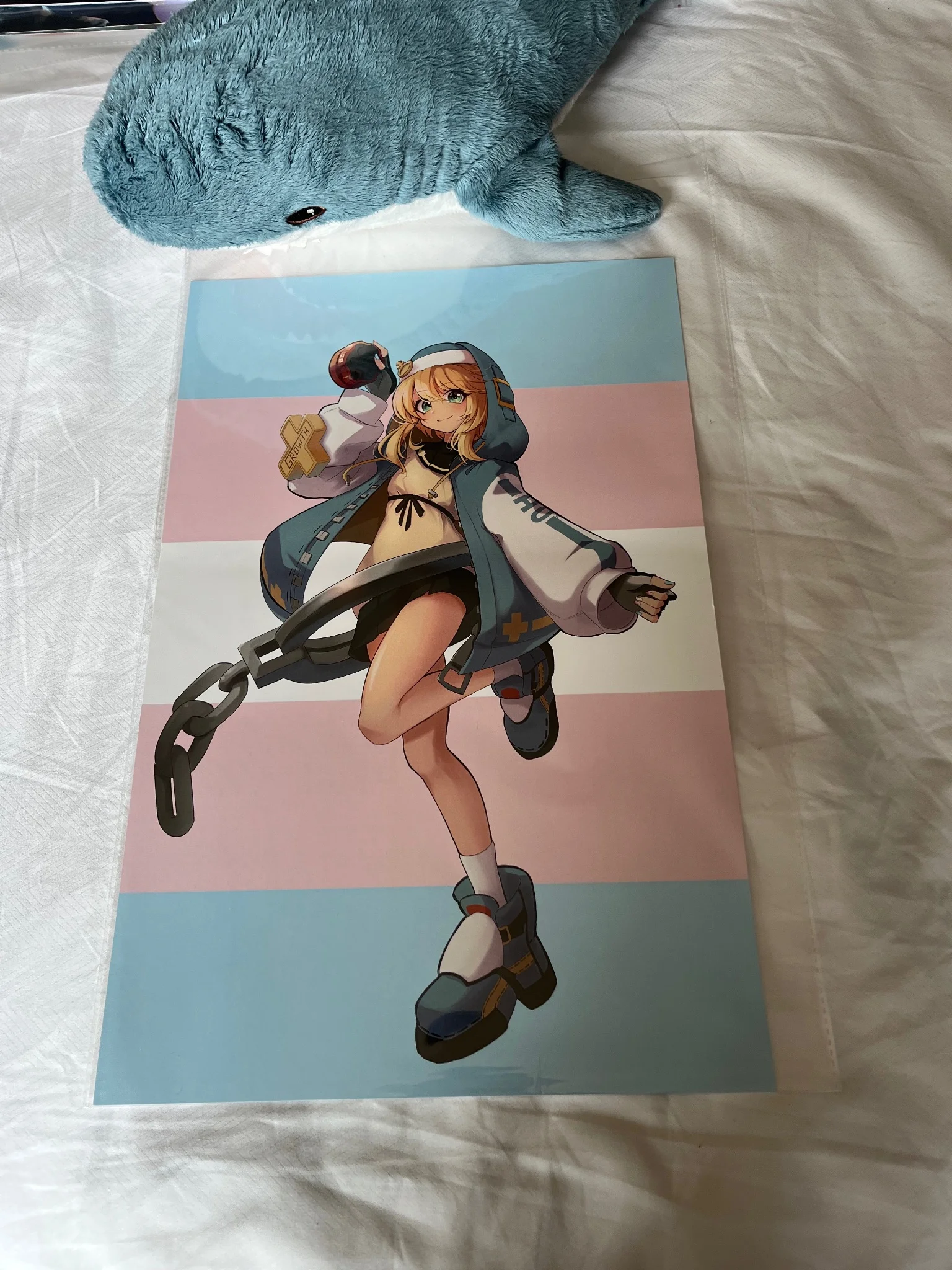 photo of a printed art piece of bridget from guilty gear, with the background of the print being the trans pride flag. above the print in the background is a mini blahaj