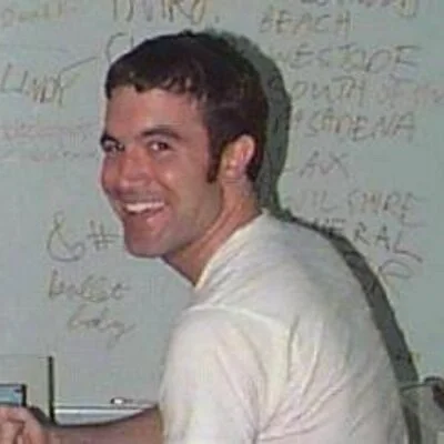the classic Myspace Tom profile picture in his white shirt, sitting in front of a whiteboard, holding a writing utensil while smilimg 