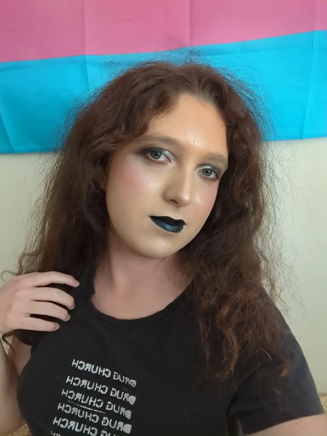 pale transfem wears goth makeup consisting of black and silver eyeshadow and black lipstick. it is also wearing a black shirt with pixel art of Saturn devouring his child