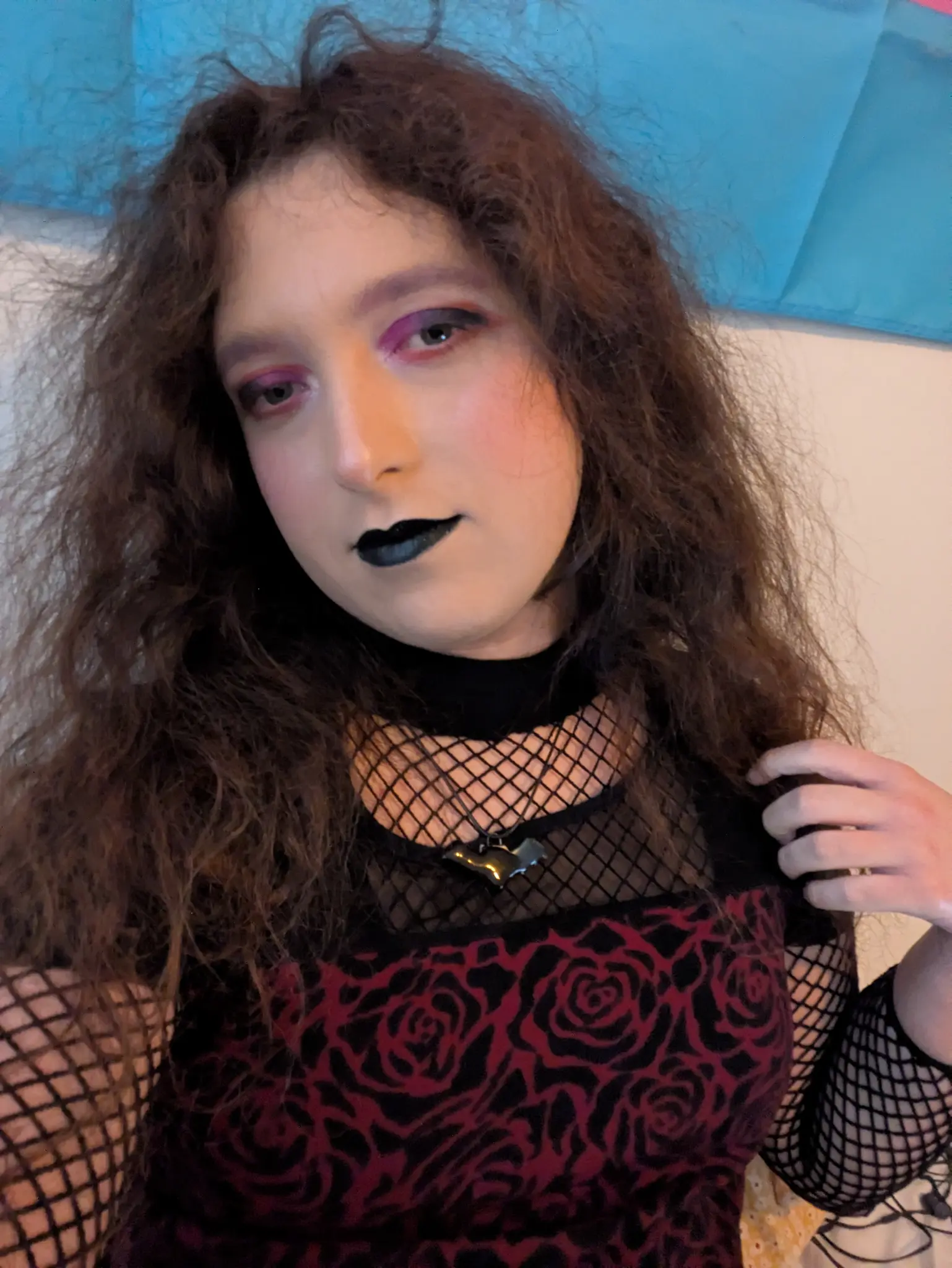 pale transfem wears red and black makeup, a fishnet shirt, and a rose shirt with a mesh shoulder region, and a bat necklace