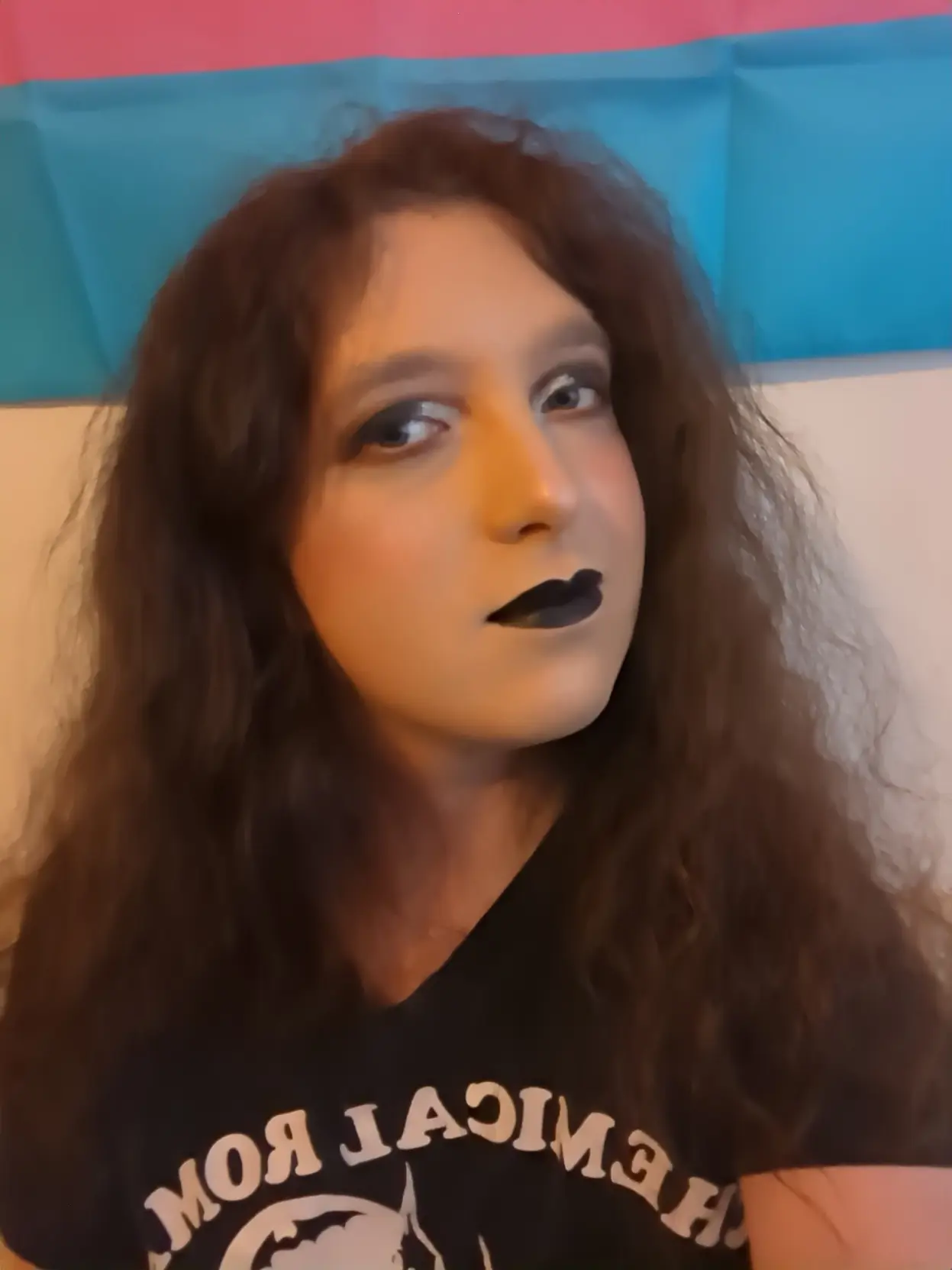 pale transfem wears goth makeup consisting of black and silver eyeshadow and black lipstick. it is also wearing a black shirt with pixel art of Saturn devouring his child