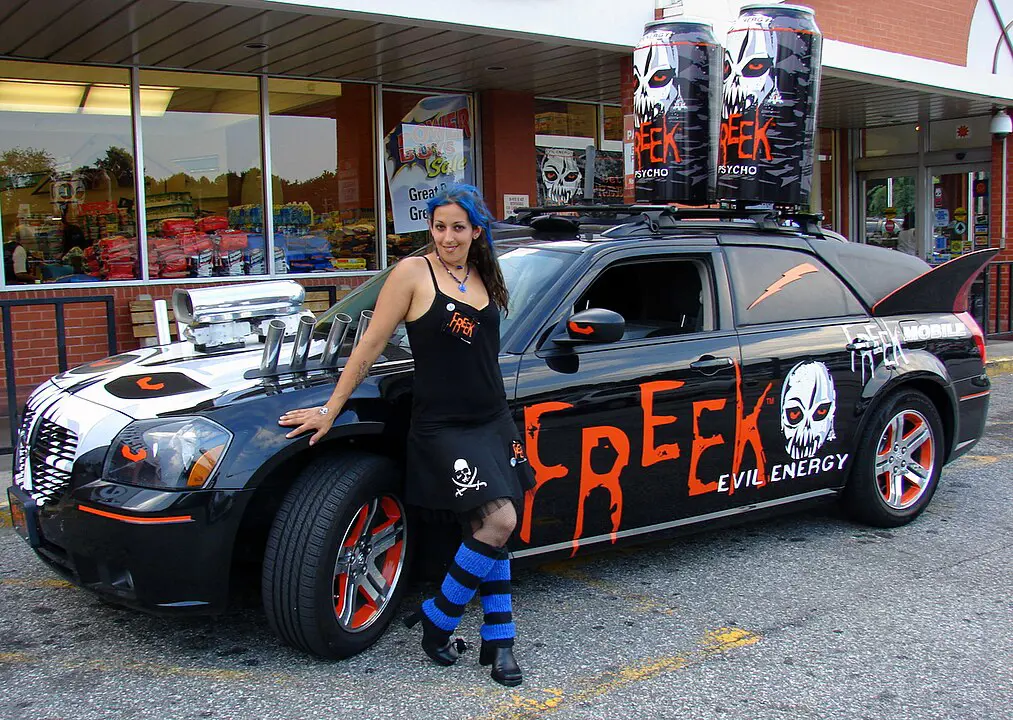 a woman in mid 2000s emo fashion stands in front of a car decked out in advertisement for a now defunct energy drink called Freek. The car is black and has two fake smoke stacks that look like energy drink cans