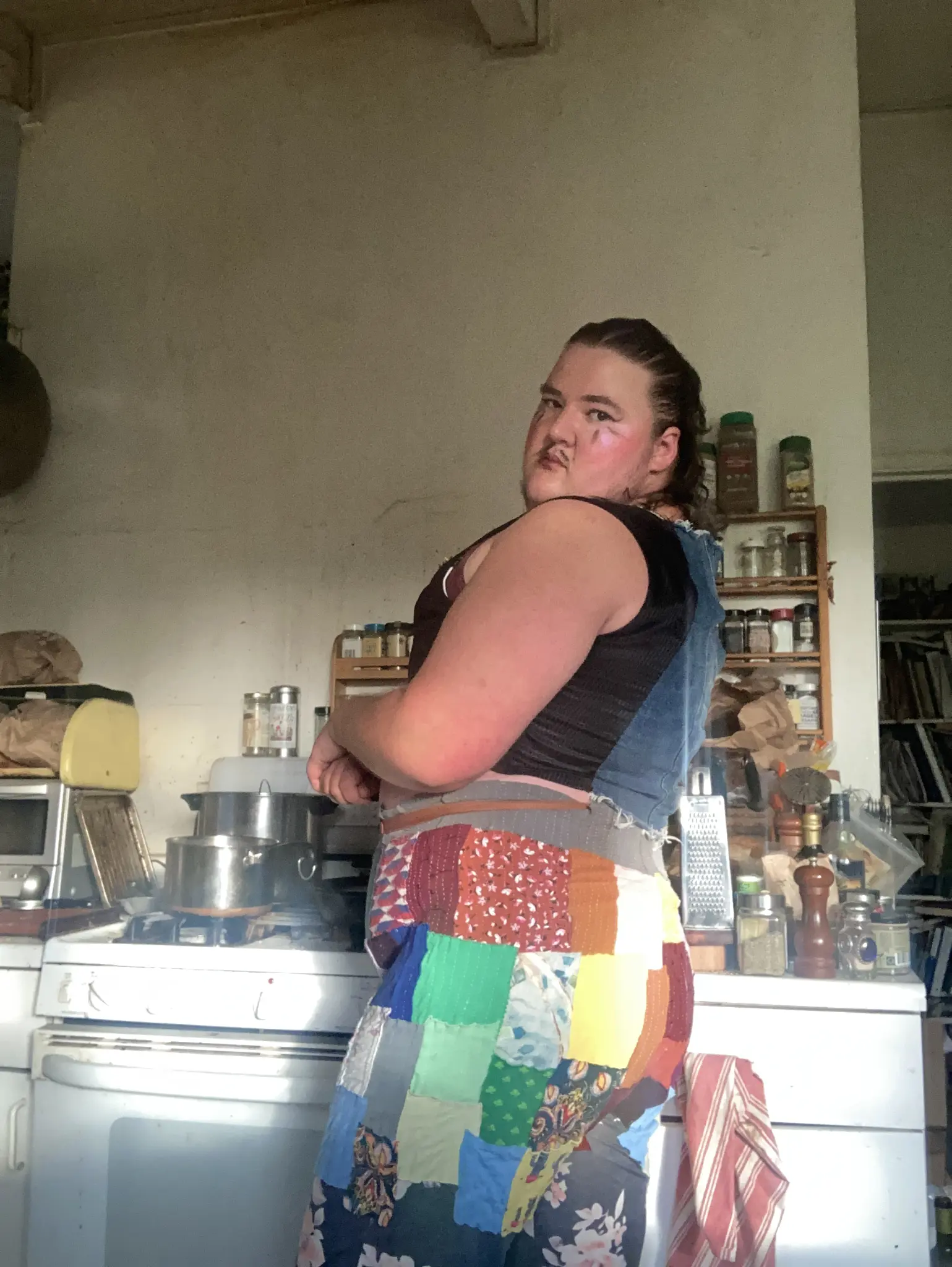 Me wearing patchwork pants and a denim and black fabric vest in front of a kitchen stove