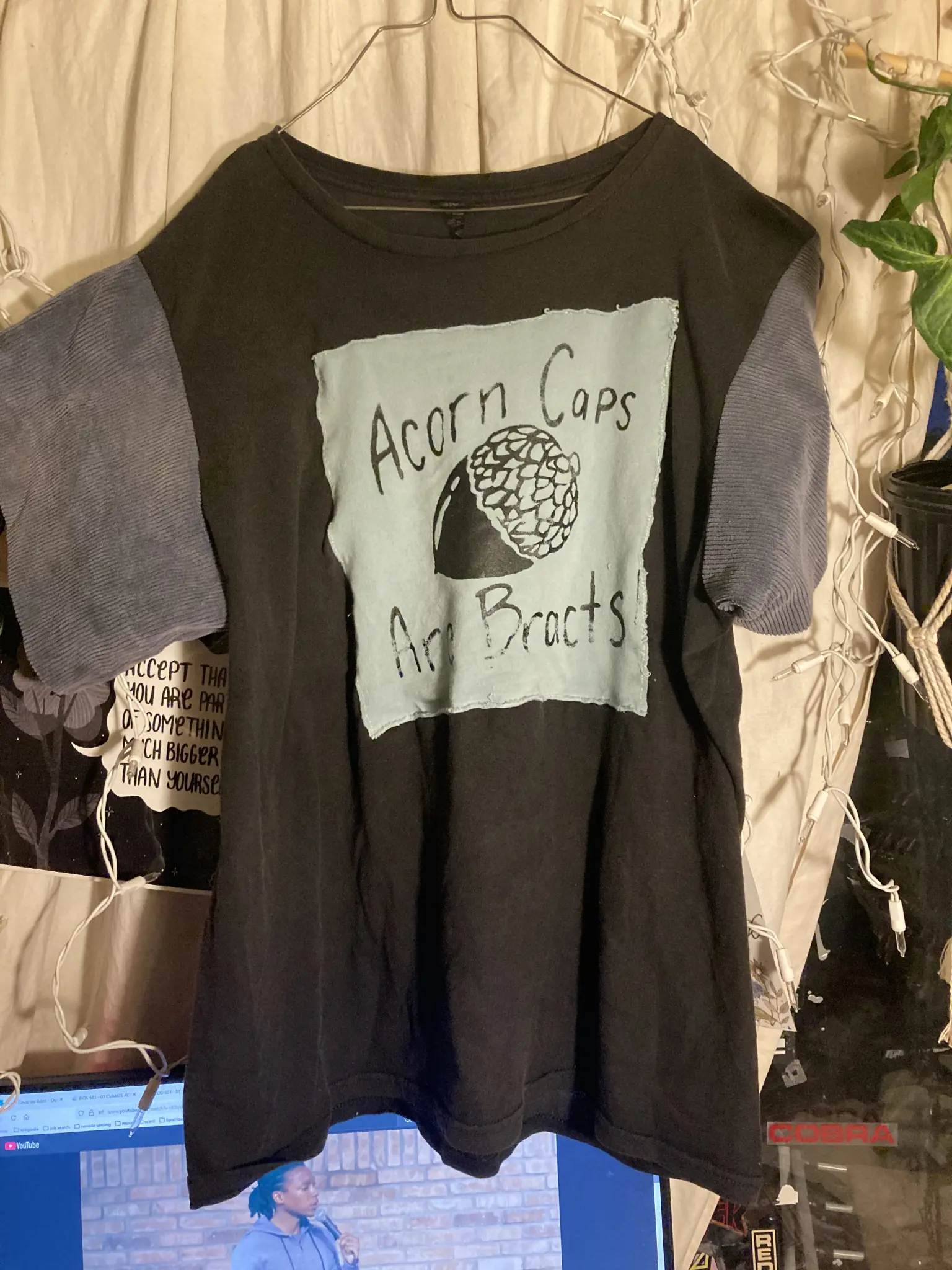 black t shirt with blue sleeves and a blue patch that says "acorn caps are bracts!" with a image of an acorn. hanging in front of a white curtain