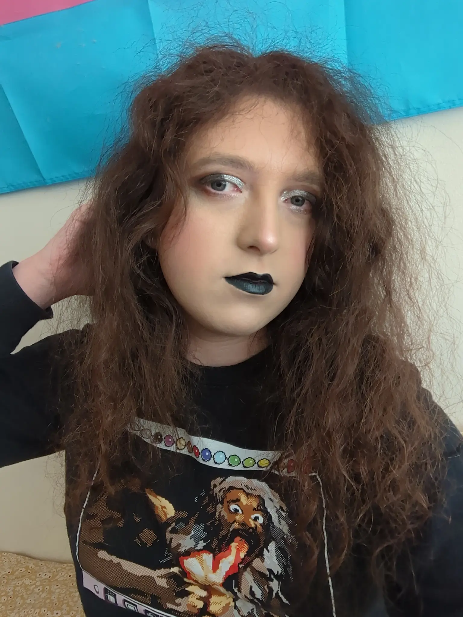 pale transfem wears goth makeup consisting of black and silver eyeshadow and black lipstick. it is also wearing a black shirt with pixel art of Saturn devouring his child