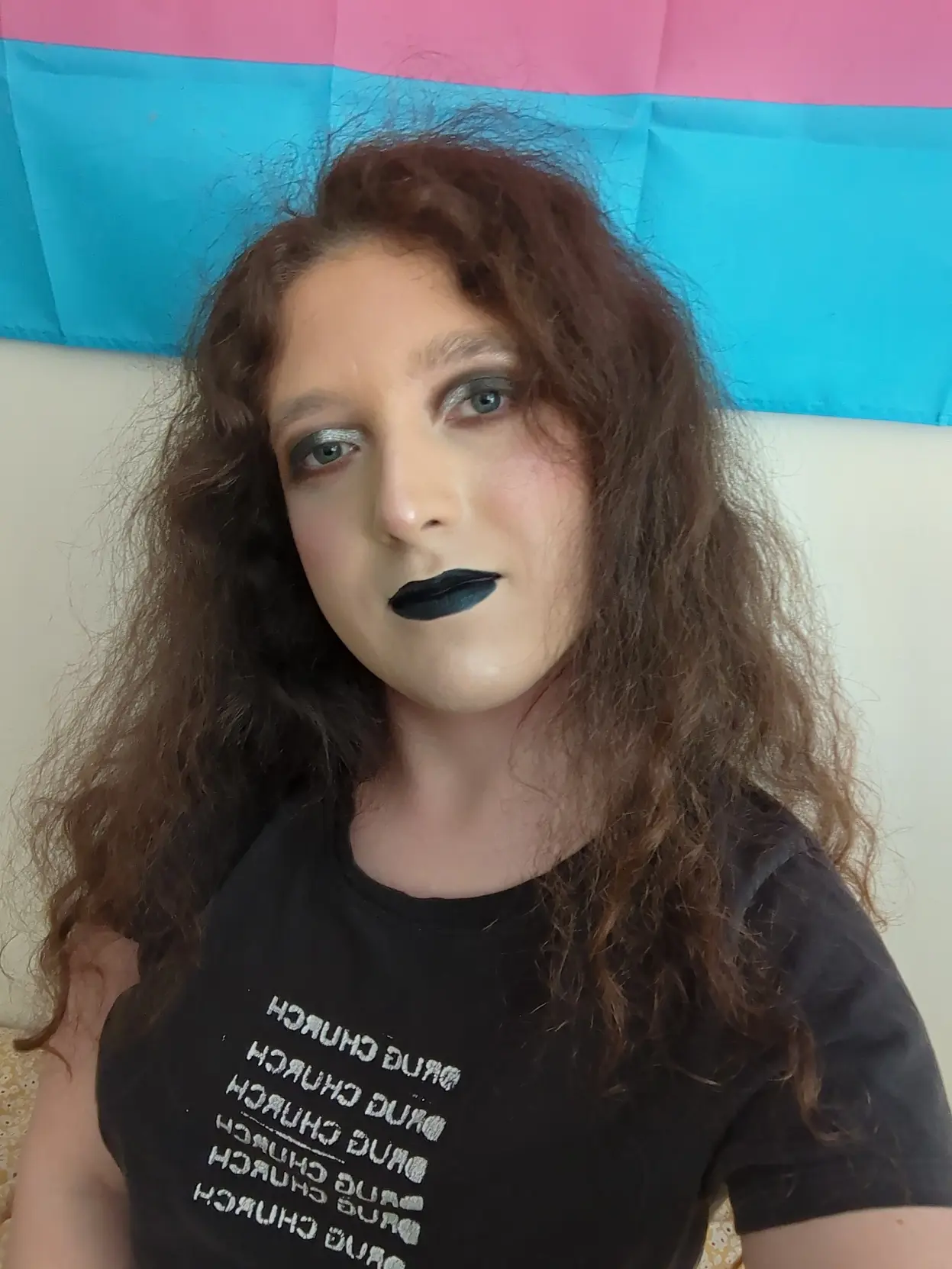 pale transfem wears goth makeup consisting of black and silver eyeshadow and black lipstick. it is also wearing a black shirt with pixel art of Saturn devouring his child
