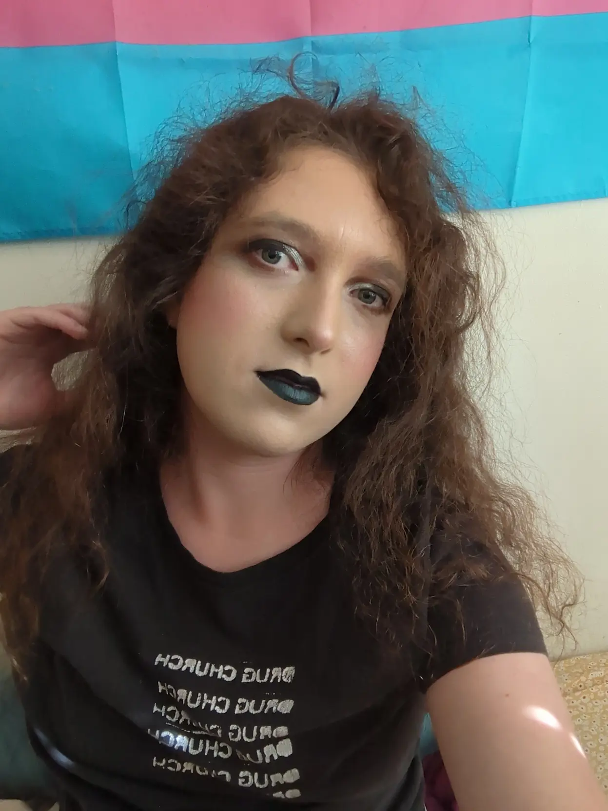 pale transfem wears goth makeup consisting of black and silver eyeshadow and black lipstick. it is also wearing a black shirt with pixel art of Saturn devouring his child